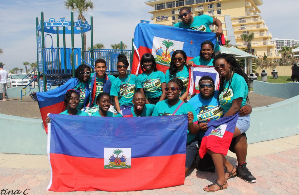 Group of young Haitian-Americans holding national flags and wearing matching shirts at a cultural celebration in a park.