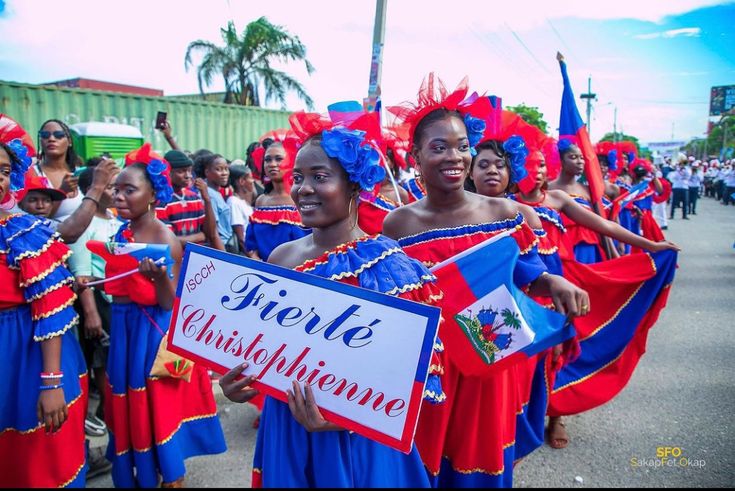 Group of Haitian women in traditional red and blue dresses parading with a "Fierté Christophienne" sign and national flags.