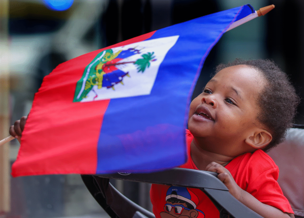 Smiling toddler holding and looking up at a waving Haitian flag during a celebration.